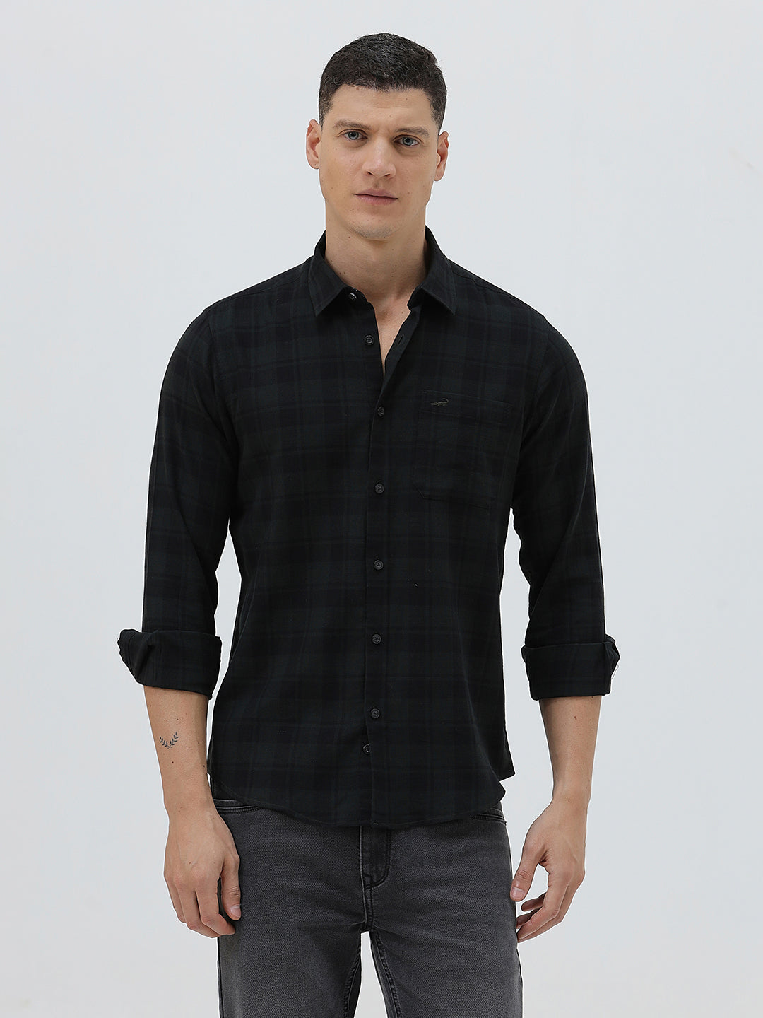 A man wearing a black long-sleeve button-up shirt with a subtle dark plaid pattern, paired with dark gray jeans, standing against a light gray studio background.