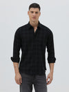 A man wearing a black long-sleeve button-up shirt with a subtle dark plaid pattern, paired with dark gray jeans, standing against a light gray studio background.