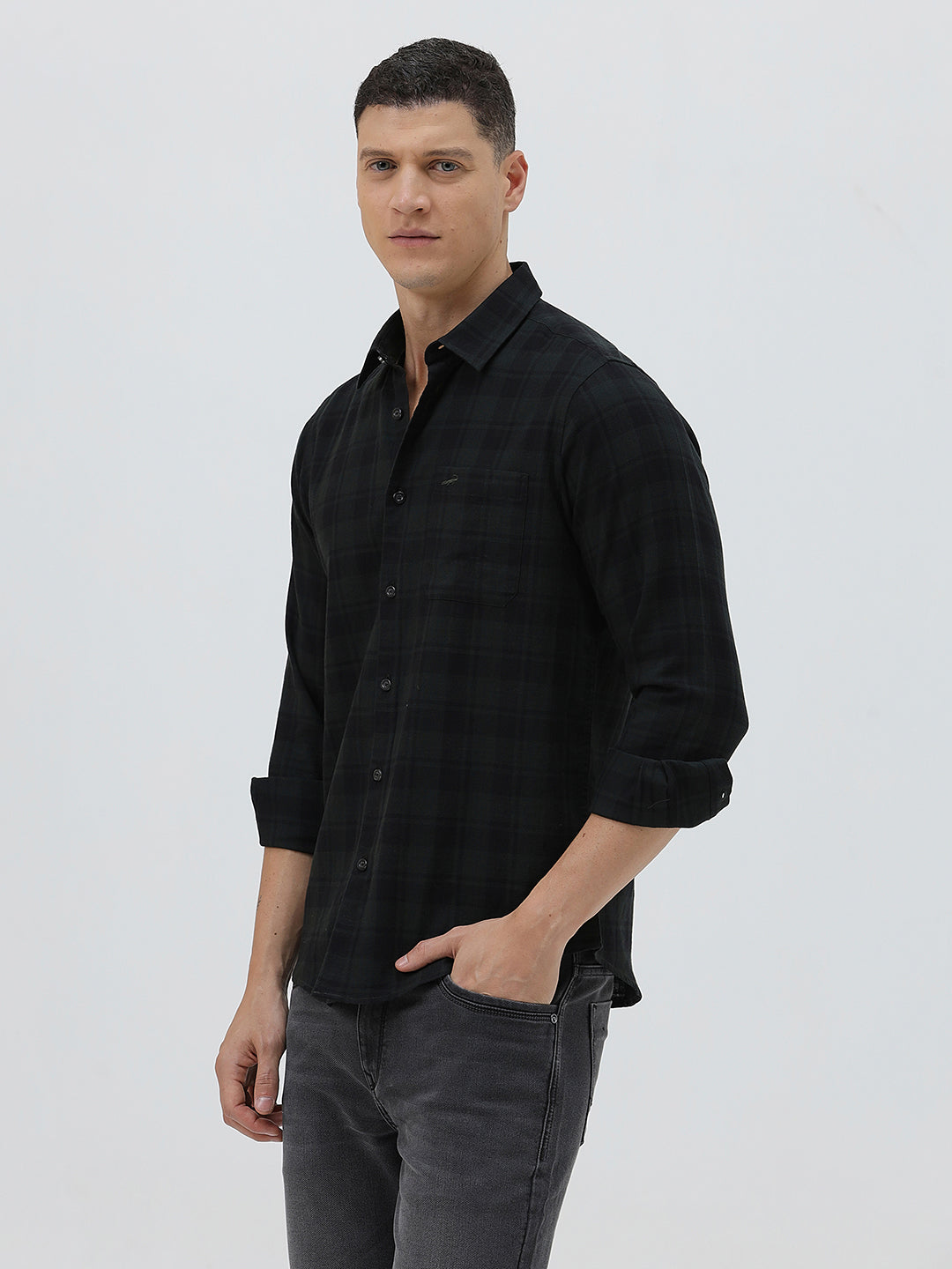A man wearing a black long-sleeve button-up shirt with a subtle dark plaid pattern, paired with dark gray jeans, standing against a light gray studio background.