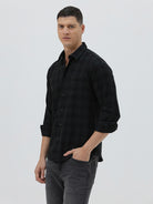 A man wearing a black long-sleeve button-up shirt with a subtle dark plaid pattern, paired with dark gray jeans, standing against a light gray studio background.