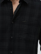 A man wearing a black long-sleeve button-up shirt with a subtle dark plaid pattern, paired with dark gray jeans, standing against a light gray studio background.