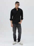 A man wearing a black long-sleeve button-up shirt with a subtle dark plaid pattern, paired with dark gray jeans, standing against a light gray studio background.