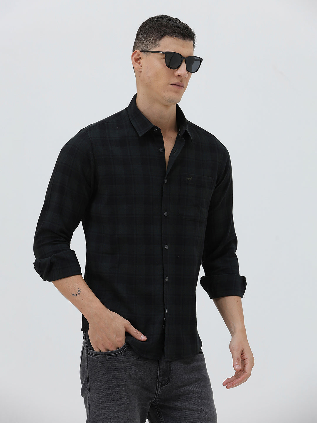 A man wearing a black long-sleeve button-up shirt with a subtle dark plaid pattern, paired with dark gray jeans, standing against a light gray studio background.
