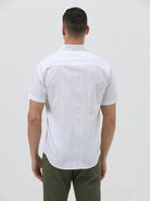 A man wearing a white short-sleeve button-up shirt with subtle vertical stripes and olive green trousers, standing against a plain light gray background.