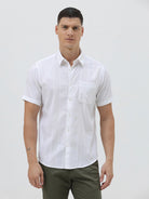 A man wearing a white short-sleeve button-up shirt with subtle vertical stripes and olive green trousers, standing against a plain light gray background.