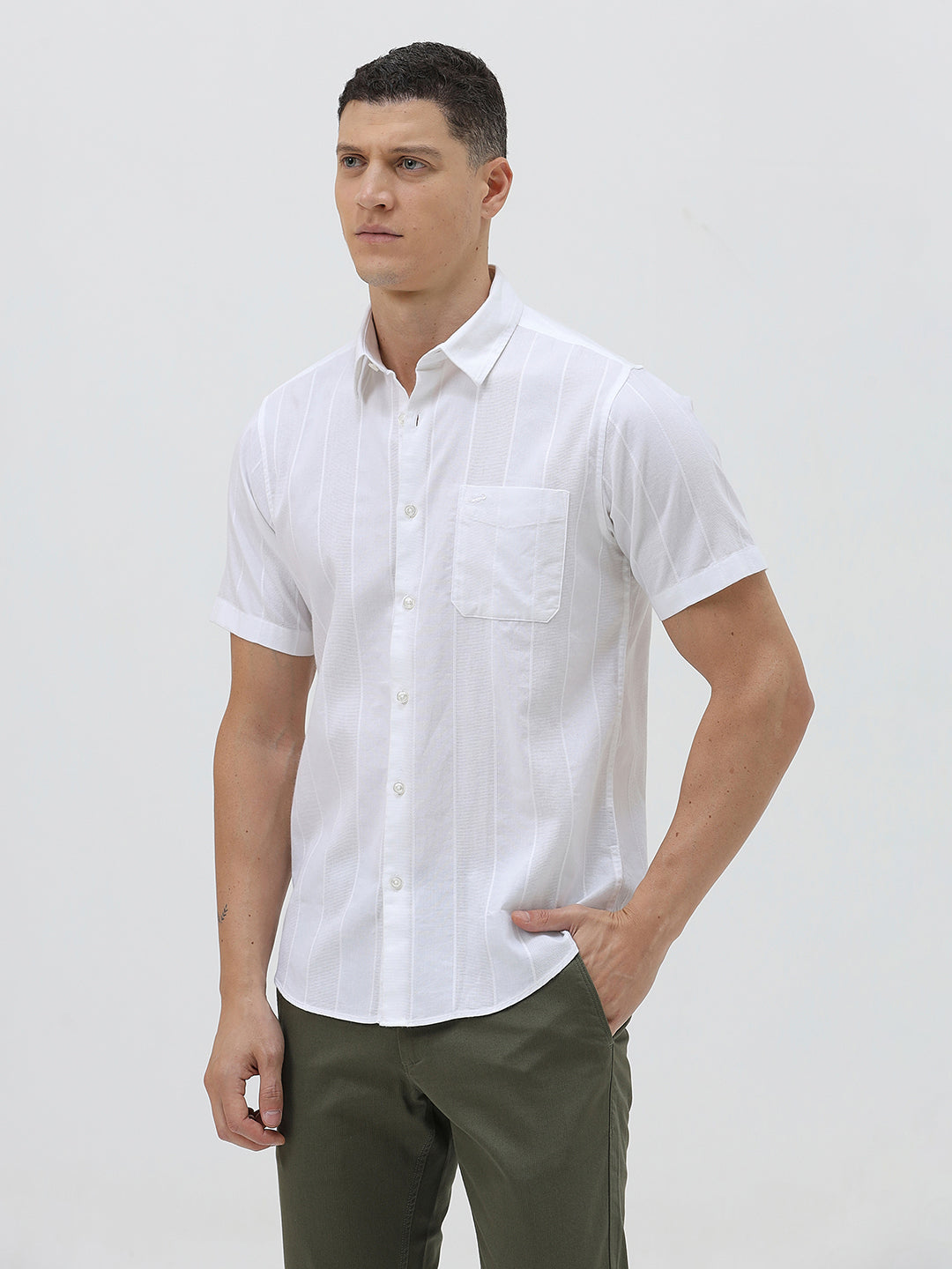 A man wearing a white short-sleeve button-up shirt with subtle vertical stripes and olive green trousers, standing against a plain light gray background.