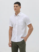 A man wearing a white short-sleeve button-up shirt with subtle vertical stripes and olive green trousers, standing against a plain light gray background.