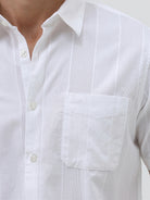 A man wearing a white short-sleeve button-up shirt with subtle vertical stripes and olive green trousers, standing against a plain light gray background.