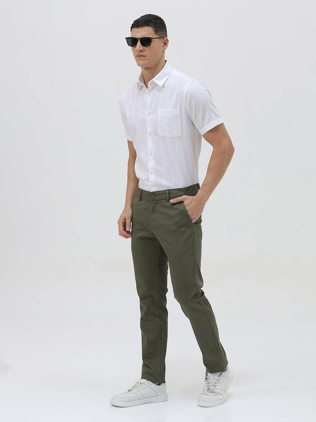 A man wearing a white short-sleeve button-up shirt with subtle vertical stripes and olive green trousers, standing against a plain light gray background.