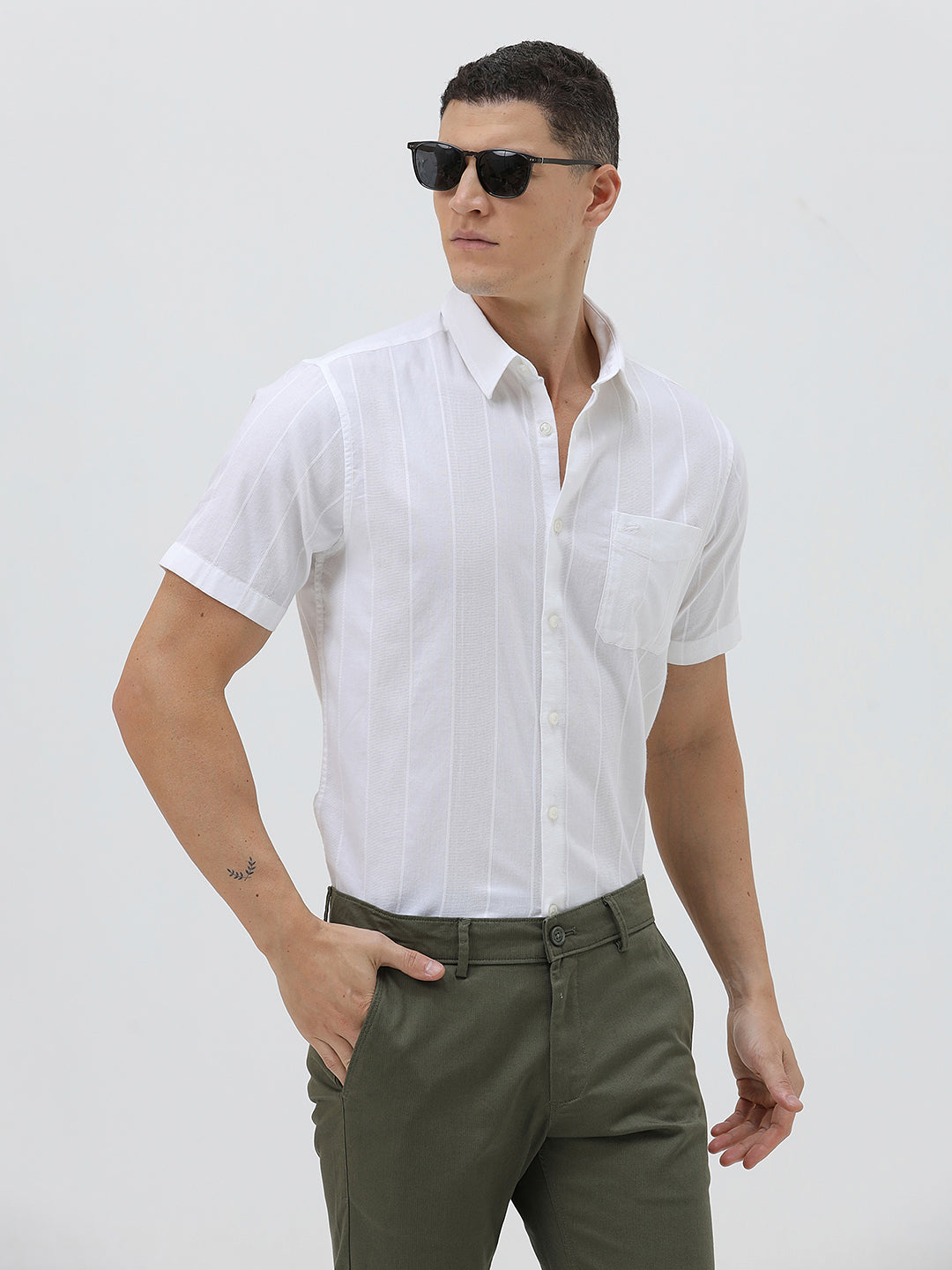 A man wearing a white short-sleeve button-up shirt with subtle vertical stripes and olive green trousers, standing against a plain light gray background.