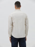 A man wearing a light beige linen long-sleeve button-up shirt with a chest pocket, paired with dark blue jeans, standing against a plain white studio background.