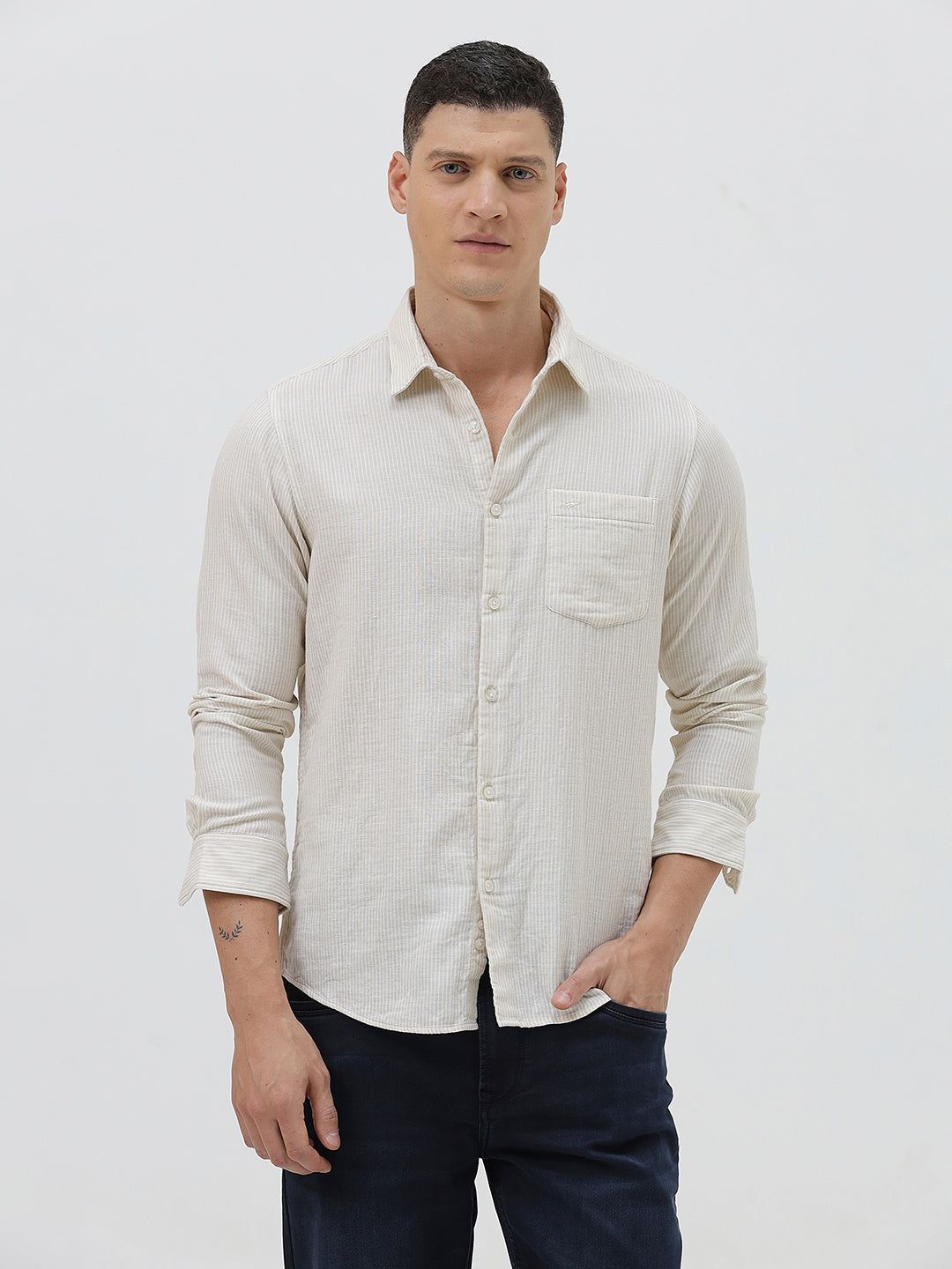 A man wearing a light beige linen long-sleeve button-up shirt with a chest pocket, paired with dark blue jeans, standing against a plain white studio background.