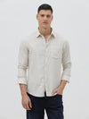 A man wearing a light beige linen long-sleeve button-up shirt with a chest pocket, paired with dark blue jeans, standing against a plain white studio background.
