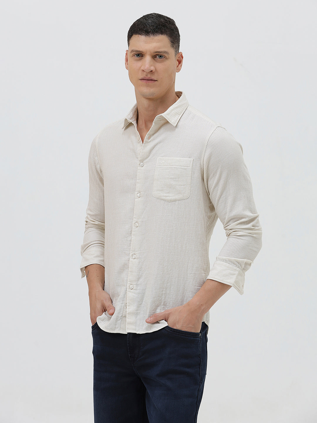 A man wearing a light beige linen long-sleeve button-up shirt with a chest pocket, paired with dark blue jeans, standing against a plain white studio background.