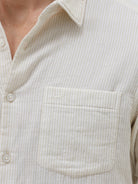 A man wearing a light beige linen long-sleeve button-up shirt with a chest pocket, paired with dark blue jeans, standing against a plain white studio background.