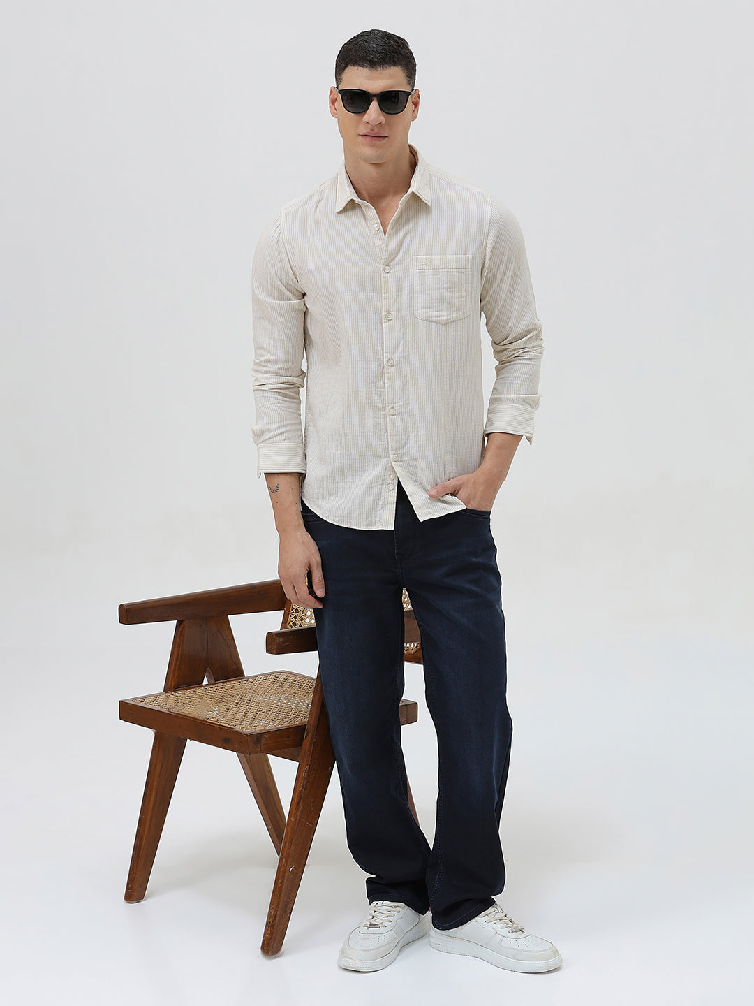 A man wearing a light beige linen long-sleeve button-up shirt with a chest pocket, paired with dark blue jeans, standing against a plain white studio background.