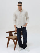 A man wearing a light beige linen long-sleeve button-up shirt with a chest pocket, paired with dark blue jeans, standing against a plain white studio background.