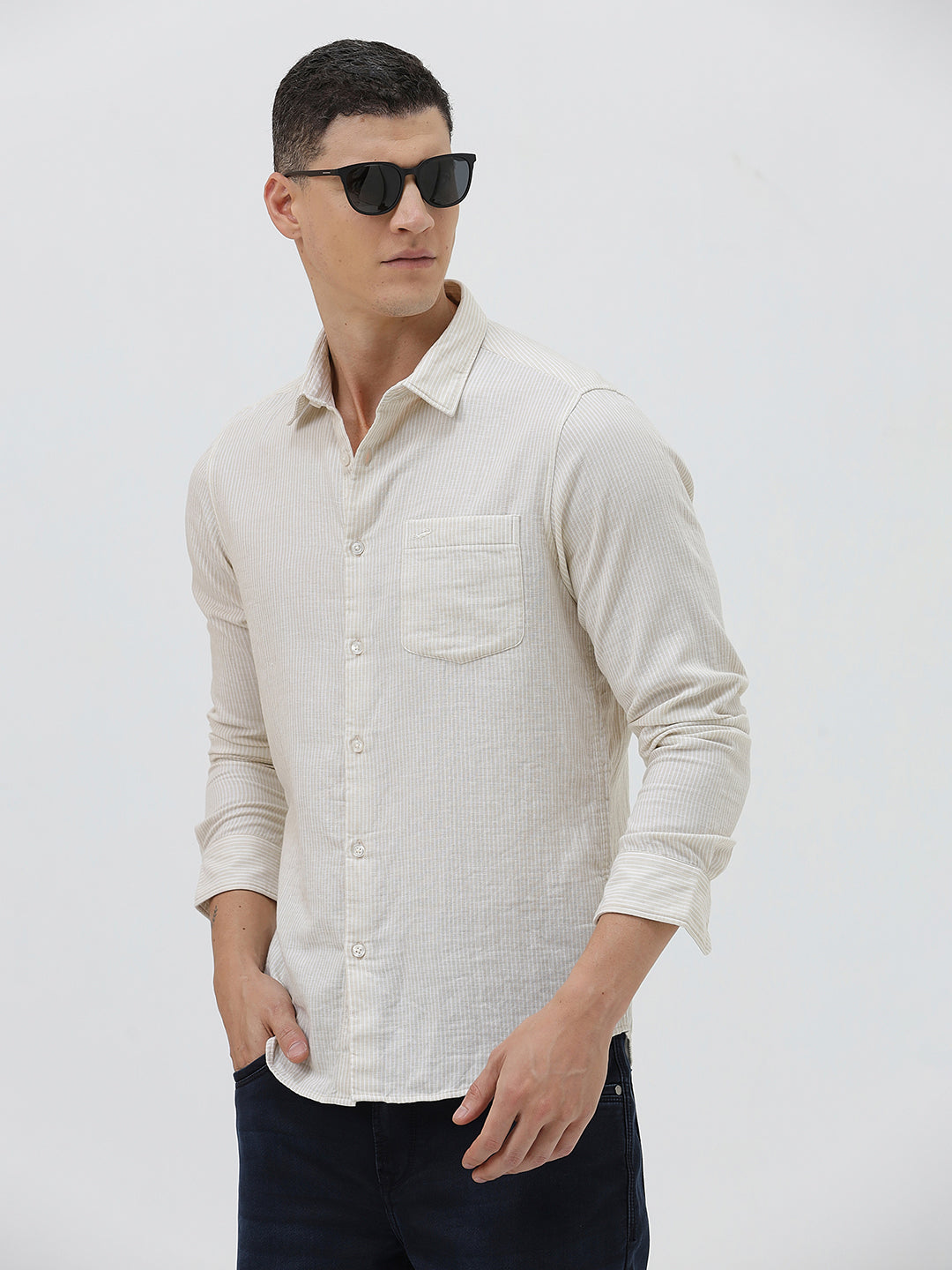 A man wearing a light beige linen long-sleeve button-up shirt with a chest pocket, paired with dark blue jeans, standing against a plain white studio background.