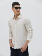 A man wearing a light beige linen long-sleeve button-up shirt with a chest pocket, paired with dark blue jeans, standing against a plain white studio background.