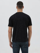 A man wearing a black short-sleeve t-shirt with a subtle tonal geometric pattern and a small 'Gildan' logo on the left chest, paired with dark blue jeans, against a light gray studio background.