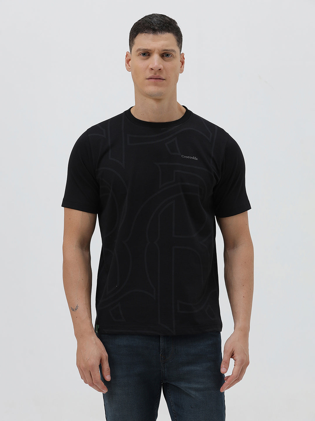 A man wearing a black short-sleeve t-shirt with a subtle tonal geometric pattern and a small 'Gildan' logo on the left chest, paired with dark blue jeans, against a light gray studio background.