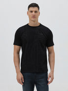 A man wearing a black short-sleeve t-shirt with a subtle tonal geometric pattern and a small 'Gildan' logo on the left chest, paired with dark blue jeans, against a light gray studio background.