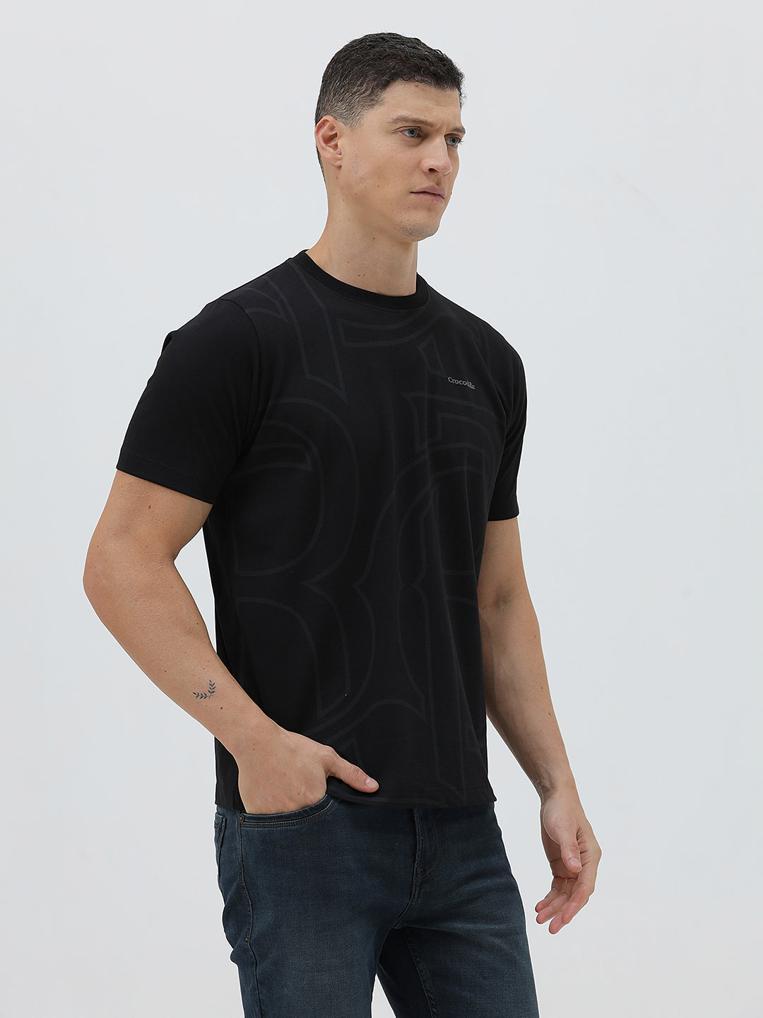 A man wearing a black short-sleeve t-shirt with a subtle tonal geometric pattern and a small 'Gildan' logo on the left chest, paired with dark blue jeans, against a light gray studio background.