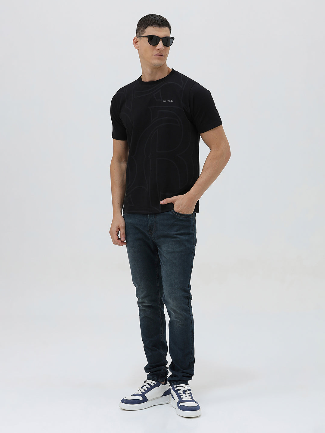A man wearing a black short-sleeve t-shirt with a subtle tonal geometric pattern and a small 'Gildan' logo on the left chest, paired with dark blue jeans, against a light gray studio background.