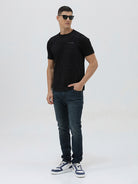 A man wearing a black short-sleeve t-shirt with a subtle tonal geometric pattern and a small 'Gildan' logo on the left chest, paired with dark blue jeans, against a light gray studio background.