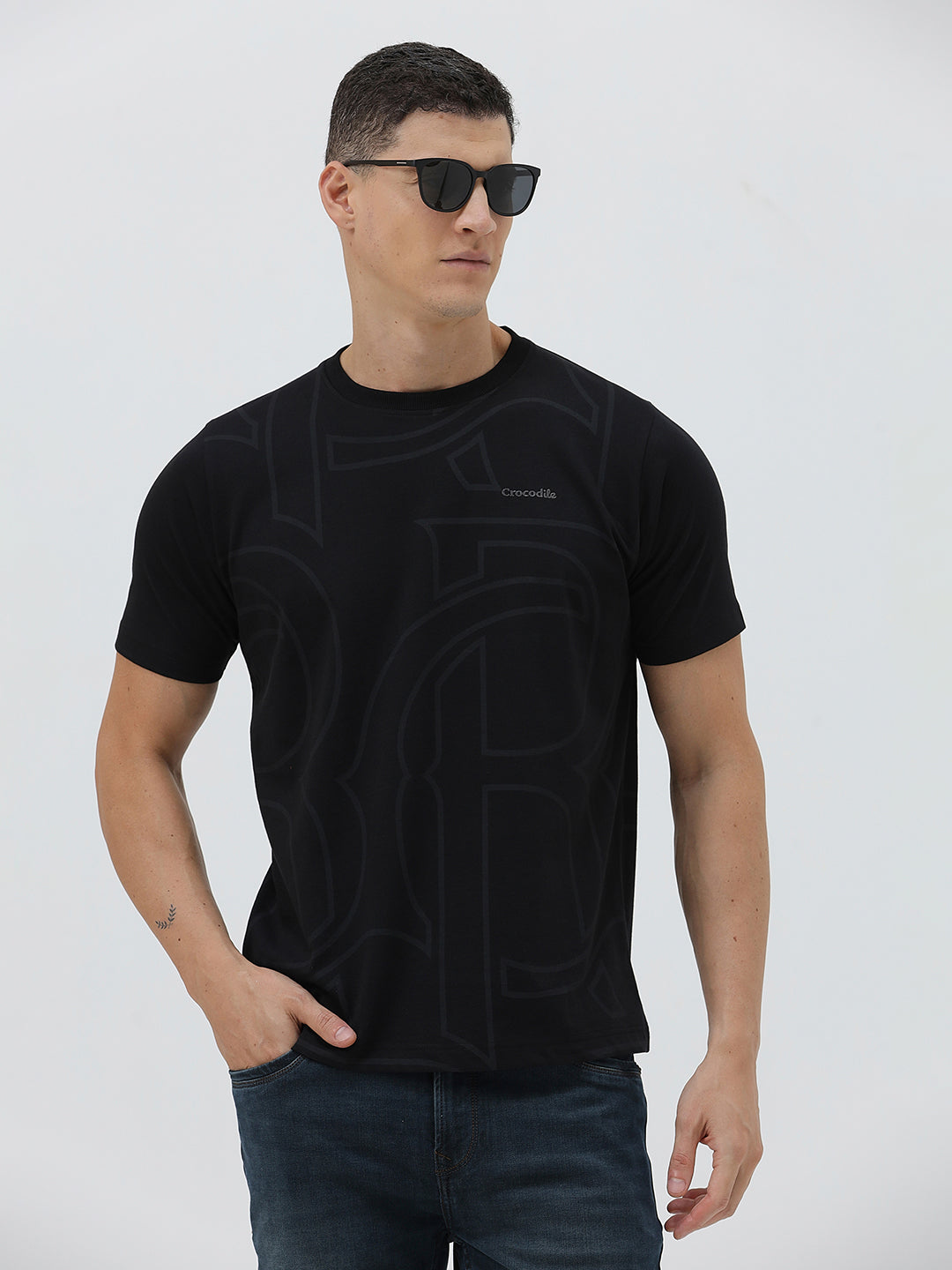 A man wearing a black short-sleeve t-shirt with a subtle tonal geometric pattern and a small 'Gildan' logo on the left chest, paired with dark blue jeans, against a light gray studio background.