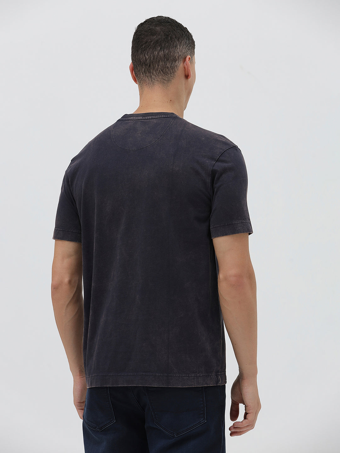 A man wearing a dark charcoal gray washed t-shirt with a blue graphic text print reading 'CROCODILE' on the chest, paired with dark blue jeans, against a light gray studio background.