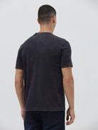 A man wearing a dark charcoal gray washed t-shirt with a blue graphic text print reading 'CROCODILE' on the chest, paired with dark blue jeans, against a light gray studio background.