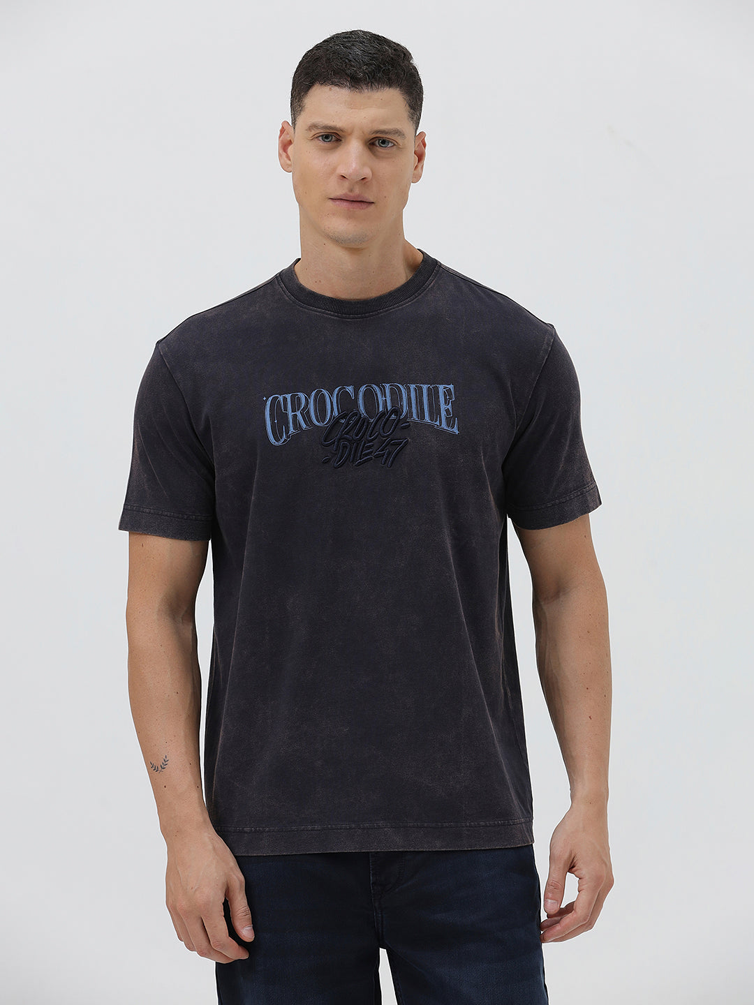 A man wearing a dark charcoal gray washed t-shirt with a blue graphic text print reading 'CROCODILE' on the chest, paired with dark blue jeans, against a light gray studio background.