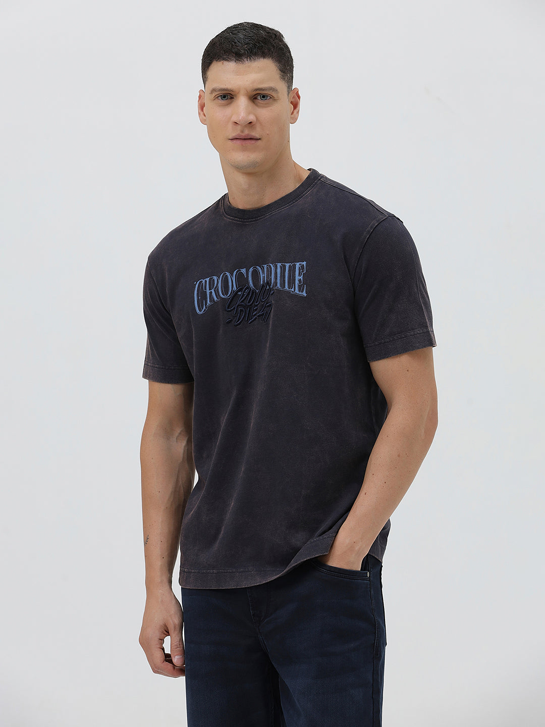 A man wearing a dark charcoal gray washed t-shirt with a blue graphic text print reading 'CROCODILE' on the chest, paired with dark blue jeans, against a light gray studio background.