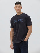 A man wearing a dark charcoal gray washed t-shirt with a blue graphic text print reading 'CROCODILE' on the chest, paired with dark blue jeans, against a light gray studio background.