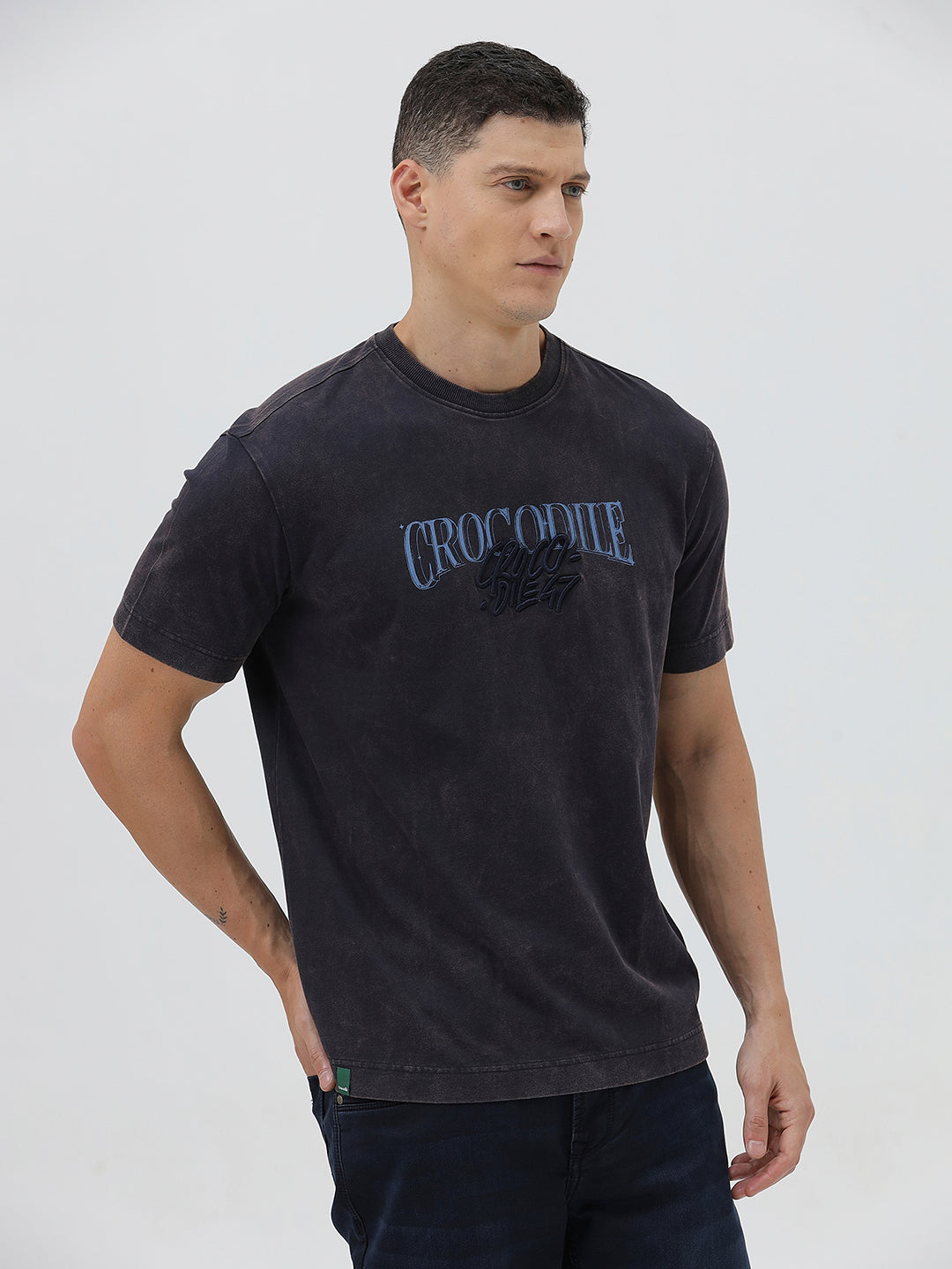 A man wearing a dark charcoal gray washed t-shirt with a blue graphic text print reading 'CROCODILE' on the chest, paired with dark blue jeans, against a light gray studio background.