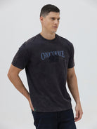 A man wearing a dark charcoal gray washed t-shirt with a blue graphic text print reading 'CROCODILE' on the chest, paired with dark blue jeans, against a light gray studio background.