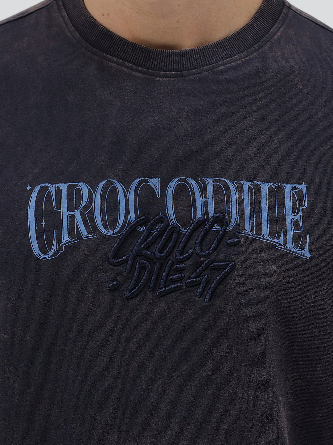 A man wearing a dark charcoal gray washed t-shirt with a blue graphic text print reading 'CROCODILE' on the chest, paired with dark blue jeans, against a light gray studio background.