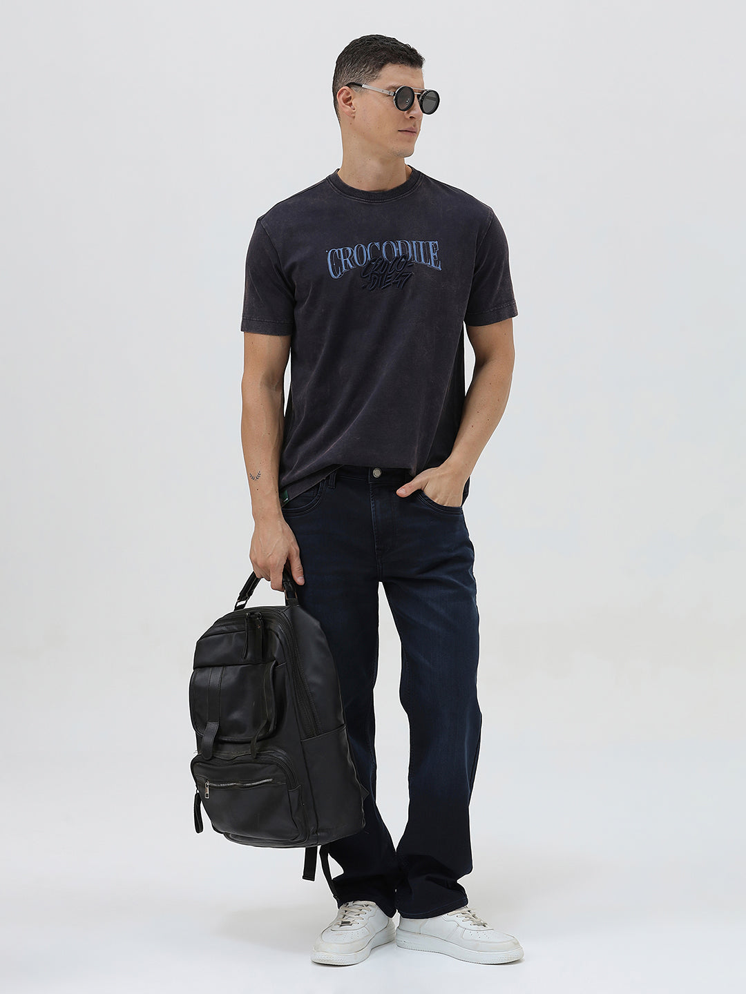 A man wearing a dark charcoal gray washed t-shirt with a blue graphic text print reading 'CROCODILE' on the chest, paired with dark blue jeans, against a light gray studio background.