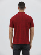 A man wearing a red short-sleeve polo shirt with a subtle tonal monogram pattern, paired with dark gray jeans, standing against a light gray studio background.