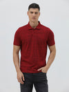 A man wearing a red short-sleeve polo shirt with a subtle tonal monogram pattern, paired with dark gray jeans, standing against a light gray studio background.