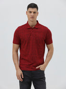 A man wearing a red short-sleeve polo shirt with a subtle tonal monogram pattern, paired with dark gray jeans, standing against a light gray studio background.