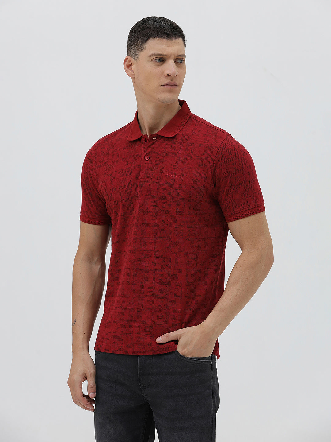 A man wearing a red short-sleeve polo shirt with a subtle tonal monogram pattern, paired with dark gray jeans, standing against a light gray studio background.