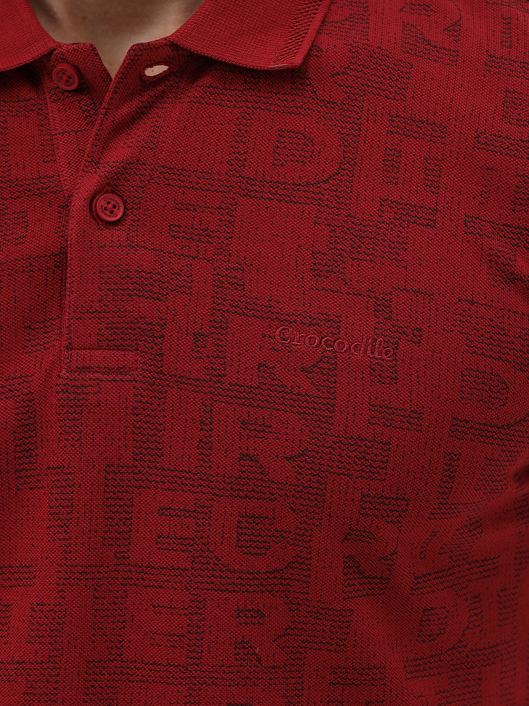 A man wearing a red short-sleeve polo shirt with a subtle tonal monogram pattern, paired with dark gray jeans, standing against a light gray studio background.