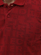 A man wearing a red short-sleeve polo shirt with a subtle tonal monogram pattern, paired with dark gray jeans, standing against a light gray studio background.