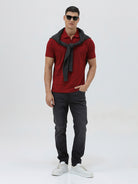 A man wearing a red short-sleeve polo shirt with a subtle tonal monogram pattern, paired with dark gray jeans, standing against a light gray studio background.