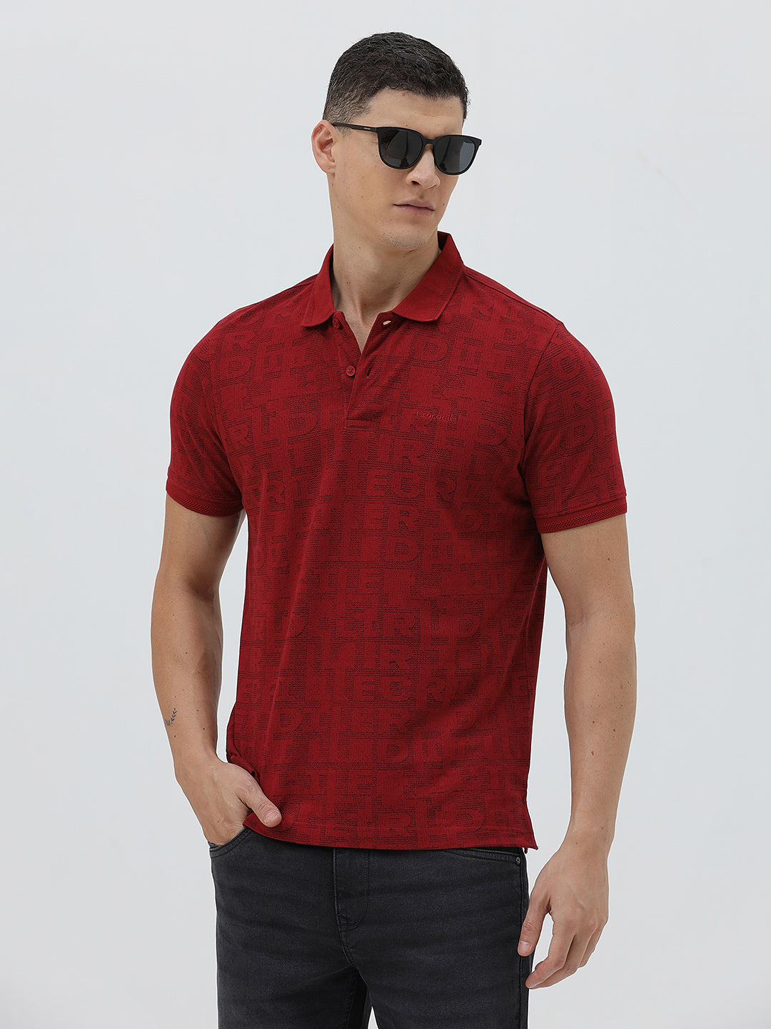 A man wearing a red short-sleeve polo shirt with a subtle tonal monogram pattern, paired with dark gray jeans, standing against a light gray studio background.
