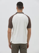 A man wearing a short-sleeve raglan polo shirt with a white body and brown sleeves, featuring a striped collar and a small 'CR' logo patch on the chest, paired with olive green pants against a light gray background.