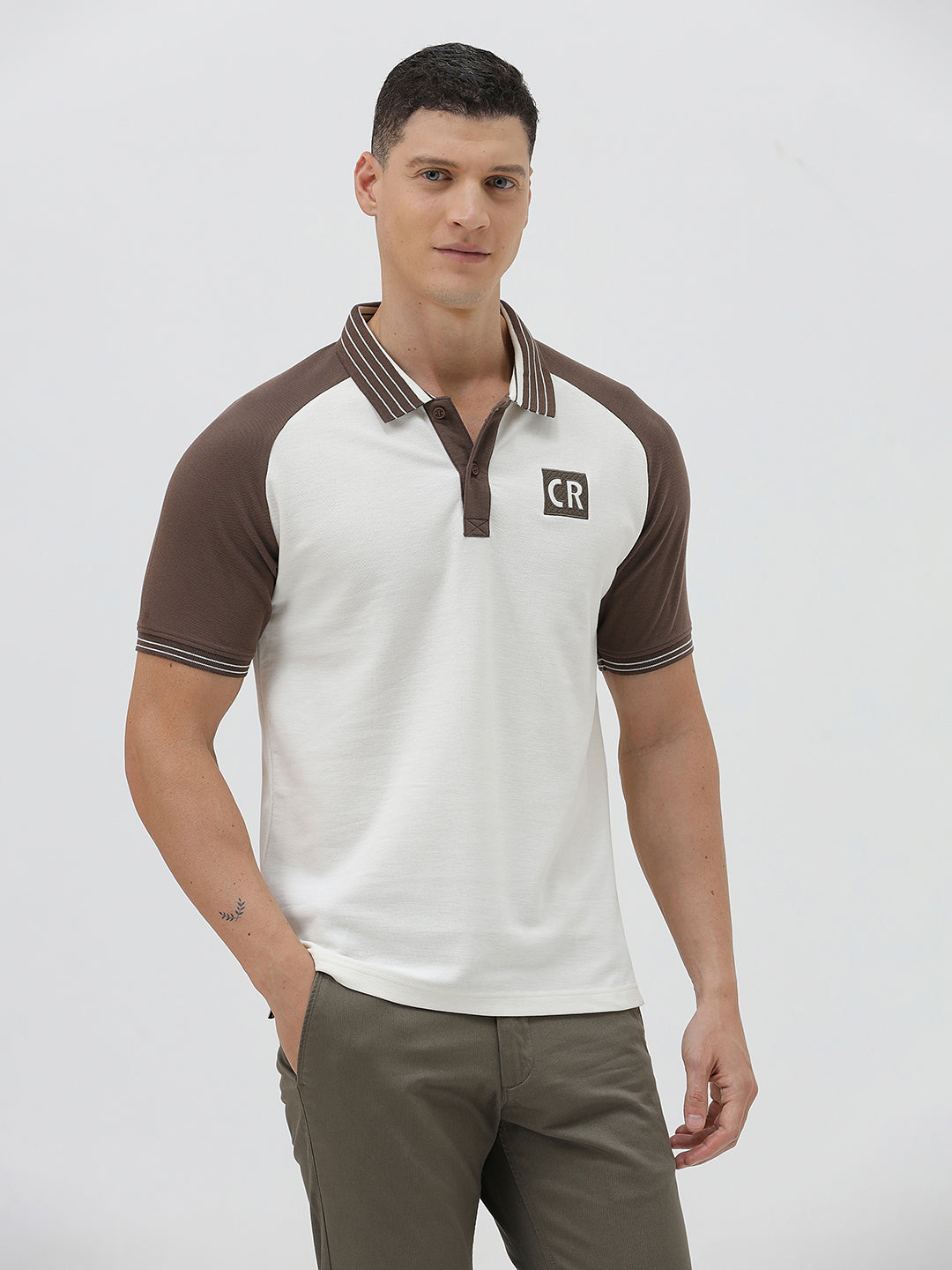 A man wearing a short-sleeve raglan polo shirt with a white body and brown sleeves, featuring a striped collar and a small 'CR' logo patch on the chest, paired with olive green pants against a light gray background.