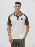 A man wearing a short-sleeve raglan polo shirt with a white body and brown sleeves, featuring a striped collar and a small 'CR' logo patch on the chest, paired with olive green pants against a light gray background.